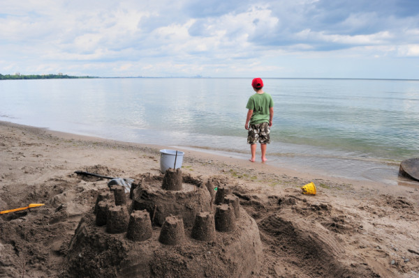 A young boy standing on the shores of Lake Ontario, looks toward the skyline of Toronto after building a sandcastle, Mississauga, Ontario, Canada.